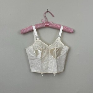 May include: Ivory-colored vintage bra with a floral lace pattern and satin-like cups. The bra has adjustable straps and is hanging on a pink padded hanger. The bra has a vintage aesthetic.