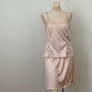 May include: A pale pink two-piece set consisting of a camisole and a skirt. The camisole features thin spaghetti straps, a straight neckline with scalloped lace trim, and a subtle tonal pattern. The matching skirt has a scalloped lace hemline.