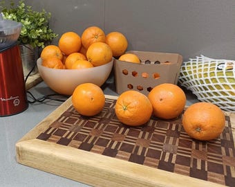 May include: A wooden cutting board with a checkered pattern in brown and light brown. There are several oranges on the board, and a few more oranges in a bowl in the background.