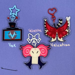May include: Three acrylic keychains on a purple background. One is a TV with the name "Vox", another is a bow with the name "Velvette", and the last is a butterfly with the name "Valentino". Each keychain has a different design and color scheme.
