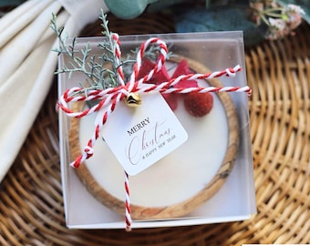 May include: A Christmas-themed gift set in a clear box. The set includes a round, white item with a wooden border, decorated with red and white twine, a sprig of greenery, and a tag that reads "Merry Christmas & Happy New Year."