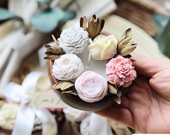 May include: A small, round wooden candle holder with a white candle in the center. The candle is surrounded by five small, white and pink wax flowers. The flowers are arranged in a circle around the candle.