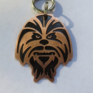 May include: A copper-colored key chain with a black silhouette of Chewbacca's face. The keychain has a silver ring.