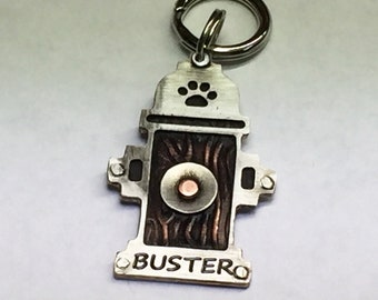 May include: Silver metal dog tag shaped like a fire hydrant with a paw print on the top, a copper colored center, and the name "Buster" engraved on the bottom.