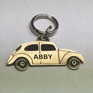 Könnte beinhalten: Ein goldfarbener Schlüsselanhänger aus Metall in Form eines Oldtimer-Volkswagen Käfers mit dem Namen "ABBY" auf der Seite graviert. Der Schlüsselanhänger hat einen silbernen Metallring.