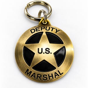 May include: A circular brass-toned metal keyring featuring a star emblem with black enamel detailing. The text DEPUTY is engraved at the top, U.S. is centred on the star, and MARSHAL is engraved at the bottom. It includes a gold-toned metal ring.