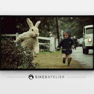 May include: A framed print featuring a large, textured rabbit sculpture next to a young child running on a path. The child wears a blue hooded jacket. A vintage car is in the background.
