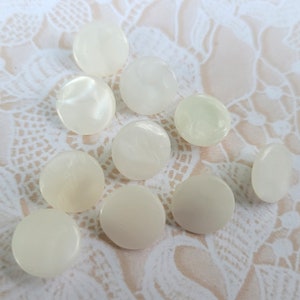 May include: A collection of ivory-colored, round buttons with a slightly pearlescent sheen. The buttons vary in tone, with some appearing more opaque and others translucent. They are scattered on a cream-colored, lace-patterned surface.