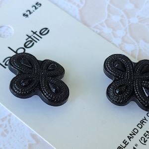 May include: Two black buttons with a four-lobed, clover-like shape and a textured, braided border design. The buttons are attached to a white display card labeled "La Petite" with the text "25MM" and "2 ON" printed on the packaging.