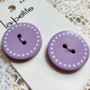 May include: Two round, lavender-colored buttons with a white dotted border and two center holes, attached to a white la petite brand display card. The buttons feature a smooth finish and are secured with thin silver-toned metal wire loops.