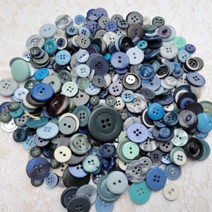 May include: A large collection of assorted sewing buttons in various shades of blue, teal, and grey. The buttons vary in size, shape, and material, featuring two-hole and four-hole designs, with some having smooth, textured, or patterned surfaces.