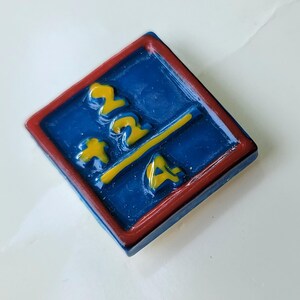 May include: A square, blue and red pin with yellow accents. The pin features the numbers "222" and the letter "A" in yellow, with a yellow line dividing the numbers and letter. The pin is set against a white background.