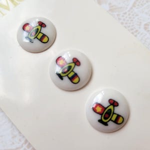 May include: Three white, round buttons with a colorful airplane design. The airplanes have red, yellow, and blue accents. The buttons are on a light cream-colored surface.