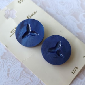 May include: Two navy blue buttons with a raised leaf design, attached to a vintage card. The buttons are round and have a glossy finish. The card is off-white with some text and a small hole.
