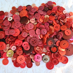 May include: A large collection of assorted sewing buttons in various shades of red, orange, and maroon. The buttons vary in size, shape, and material, featuring two or four holes, with some displaying smooth, textured, or metallic finishes.