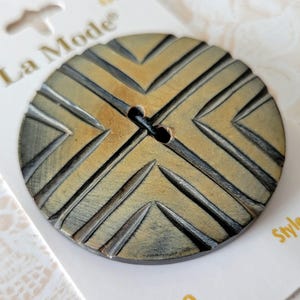 May include: A round button with a tan and dark gray chevron pattern featuring deep grooves radiating from the center. The button has two holes in the middle and is attached to a white card with the text La Mode printed in the upper left corner.