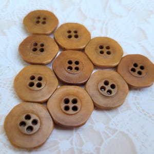May include: A collection of nine tan, octagonal-shaped buttons arranged in a triangular formation. Each button features a smooth, matte surface and a central recessed area containing four evenly spaced holes for threading.