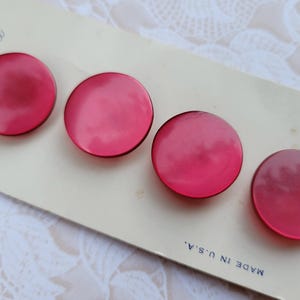 May include: Four vintage, round, bright pink buttons on a cream-colored card. The card has text that reads "Buttons" and "Made in U.S.A.". The buttons are glossy and have a slight dome shape. The card is slightly aged.