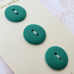 May include: Three teal-colored, oval-shaped buttons with white thread, arranged on a light beige card. The buttons have a matte finish and are evenly spaced. The card has a subtle texture.