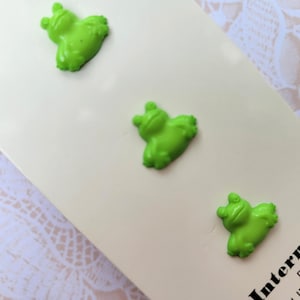 May include: Three bright green frog-shaped buttons arranged on a cream-colored card. The buttons have a three-dimensional design, with visible eyes and textured skin. The card has text that reads "International Buttons 1/4".