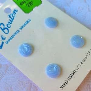 May include: A package of four light blue buttons on a cream-colored card labeled Le Bouton. The card displays the text Guaranteed Washable and Size 11mm (7/16 inches) 4 count lt blue. The buttons are round with a smooth, slightly glossy finish.
