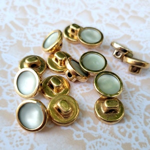 May include: A collection of 12 gold-toned buttons with white centers. The buttons are round and have a raised edge. They are arranged in a random pattern on a white background.
