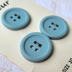 May include: Three light blue, round buttons with two holes and a metal shank. The buttons are on a cream-colored paper with text. The buttons are approximately 1.25 inches in diameter.