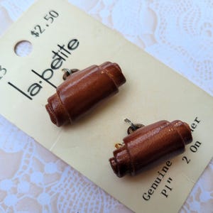 May include: Two brown leather buttons with a cylindrical design are displayed on a vintage price tag. The tag reads "la petite" and "Genuine Leather P1" 2 on". The buttons are approximately 1 inch in length.