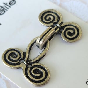 May include: A two-piece metal toggle closure with an antique brass finish, featuring a double spiral design on each side. The fastener is attached to a white display card with the text "la petite" and "CLOSURE" printed in black lettering.