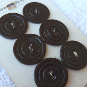 May include: Six dark brown, circular buttons with concentric circle detailing. Each button has two holes and is attached with white thread. The buttons are on a vintage paper card with faded text.