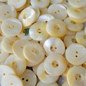 May include: A collection of round, off-white mother-of-pearl buttons, each featuring a smooth, iridescent surface and two central holes for attachment. The buttons vary slightly in their creamy, pearlescent tones and light-reflecting finish.