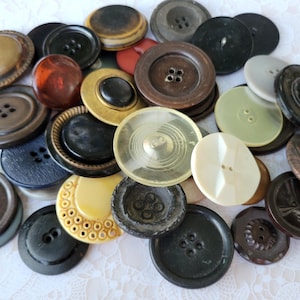 May include: A collection of assorted round buttons in various sizes, colors, and materials. The pile includes dark brown, black, cream, amber, and metallic-toned buttons with different textures, some featuring four holes, others two, and some with decorative rims.