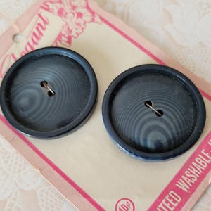 May include: Two round, dark navy blue buttons with a subtle concentric circular pattern and two center holes, attached to a beige cardboard display card featuring the word Elegant and the text Guaranteed Washable - Ironable in pink and white.