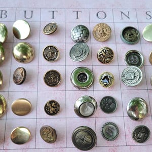 May include: Assortment of decorative buttons in various shapes, sizes, and finishes, including gold, silver, and bronze. Some buttons feature embossed designs, while others are smooth or textured. The word "BUTTONS" is printed at the top.