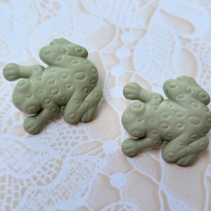 May include: Two light green frog-shaped buttons with a textured, dotted surface. The buttons are lying on a cream-colored, patterned background. The buttons are a whimsical craft supply.