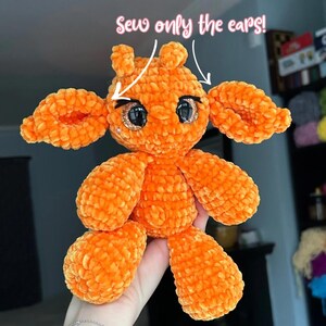 Op de afbeelding: Een oranje pluche speelgoed met grote, glinsterende ogen en een zacht, getextureerd uiterlijk. Het speelgoed heeft grote oren en ledematen, met de tekst "Sew only the ears!" erboven.