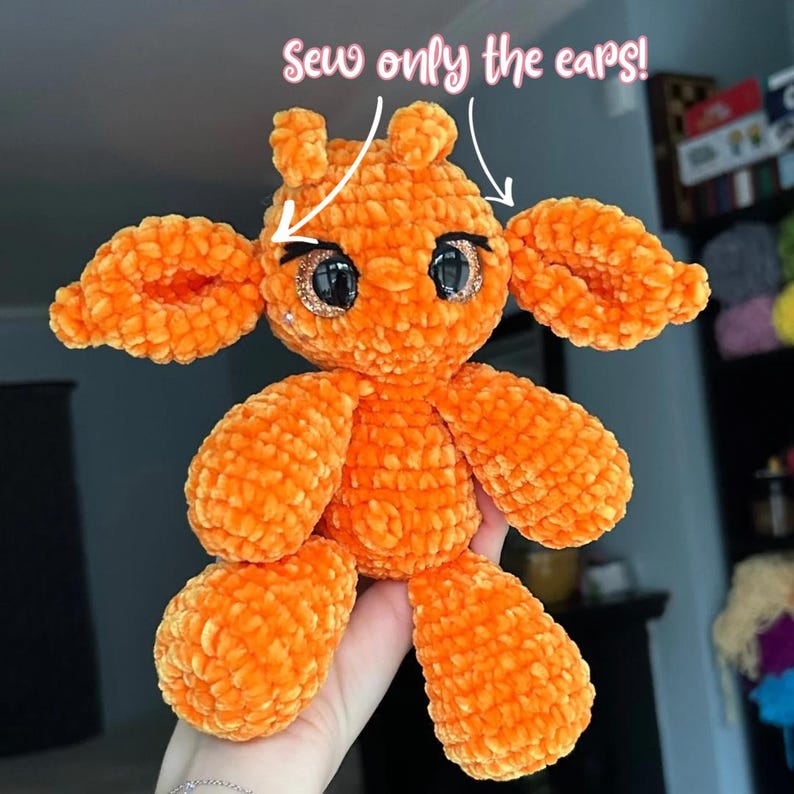 Op de afbeelding: Een oranje pluche speelgoed met grote, glinsterende ogen en een zacht, getextureerd uiterlijk. Het speelgoed heeft grote oren en ledematen, met de tekst "Sew only the ears!" erboven.