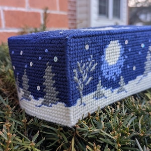 May include: A blue and white tissue box cover with a winter scene featuring trees, a moon, and snowflakes. The cover is made of fabric and has a white border.