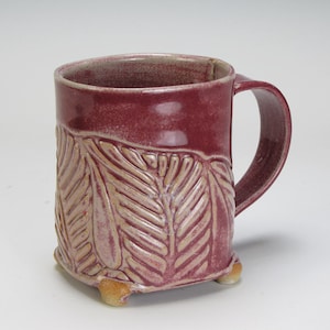 Może przedstawiać: Ceramiczny kubek z głęboką magentową glazurą i gładką górną krawędzią. Dolna część posiada teksturowany, tłoczony wzór liści paproci w jaśniejszym odcieniu. Kubek ma zakrzywione ucho i stoi na trzech małych, zaokrąglonych nóżkach.