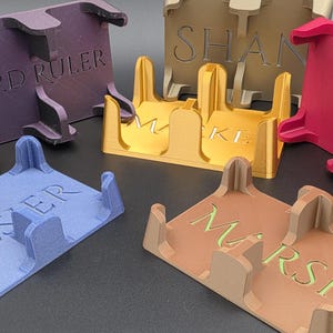 May include: A collection of colorful, 3D-printed card holders. Each holder is a different color, including purple, blue, gold, pink, and brown. The holders have text on them, such as "LORD RULER", "KELSER", "MAKE", "VIN", and "MARSH".