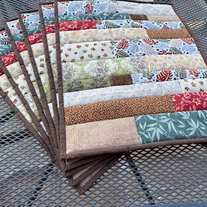 May include: A stack of handmade quilted placemats. Each placemat features a patchwork design with various fabrics in shades of red, green, brown, and cream. The edges are finished with a brown binding.