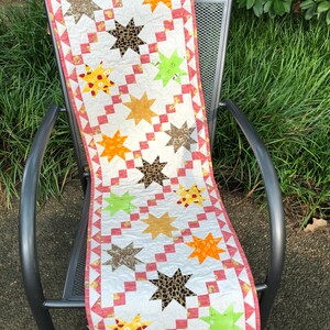 May include: A colorful star-patterned table runner. The runner features stars in various colors, including brown, orange, yellow, and green, with a leopard print star. The border is a red and white pattern. The runner is displayed on a metal chair.