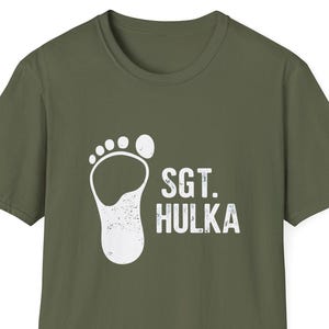 May include: Olive green t-shirt with a distressed white graphic of a footprint and the text "SGT. HULKA". The shirt has short sleeves and a crew neck. The design is centered on the front of the shirt.