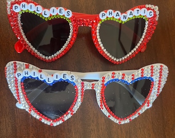 May include: Two pairs of heart-shaped sunglasses. One pair is red with the word "PHILLIES PHANATIC" spelled out in white beads. The other pair is white with red and blue stripes and the word "PHILLIES" spelled out in white beads.