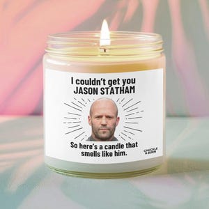 May include: A lit, clear glass jar candle with a white label featuring the text "I couldn't get you JASON STATHAM." Below the text is a photo of Jason Statham. The label also reads "So here's a candle that smells like him."