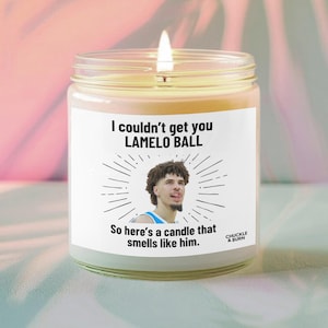 以下が含まれることがあります： 透明なガラス瓶に入った、火の灯ったキャンドル。ラベルにはバスケットボール選手の写真と「I couldn't get you Lamelo Ball. So here's a candle that smells like him.」というテキストが記載されています。