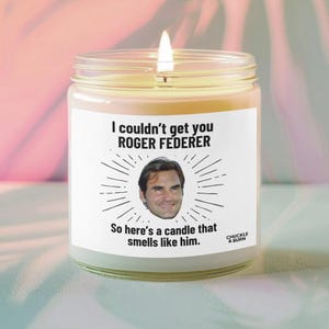 May include: A lit candle in a glass jar with a white label. The label features the text "I couldn't get you ROGER FEDERER" above a photo of Roger Federer. Below the photo, it reads "So here's a candle that smells like him."