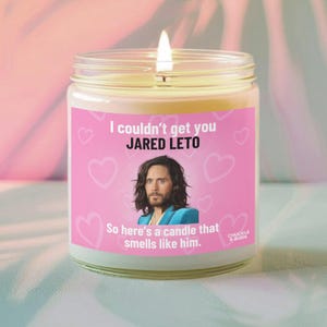May include: A lit scented candle in a glass jar with a pink label featuring the text "I couldn't get you JARED LETO" and a photo of Jared Leto. The label also reads "So here's a candle that smells like him."