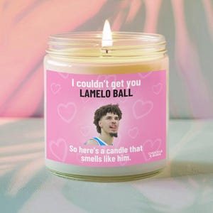 以下が含まれることがあります： ピンク色のラベルが付いた、火の灯ったガラス瓶のキャンドル。ラベルには「I couldn't get you LAMELO BALL」というテキストとバスケットボール選手の写真が掲載されています。ラベルには「So here's a candle that smells like him.」とも書かれています。