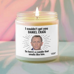 May include: A lit candle in a glass jar with a white label. The label features a photo of Daniel Craig and the text "I couldn't get you Daniel Craig. So here's a candle that smells like him."